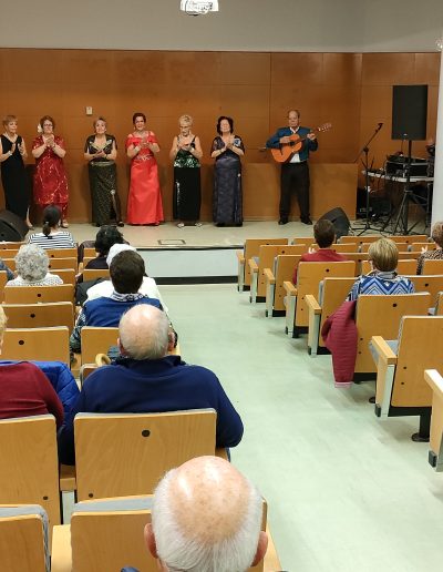 Santa Coloma de Gramenet. PlaybackMENT. View from the audience of elderly people watching a show. On stage, six women wearing long dresses clap their hands and a man plays the guitar.