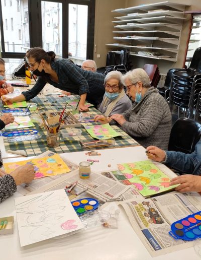 A group of elderly people sitting around a long table covered with paper, paint brushes and paint. Each person is concentrating on their own creation. A younger lady bends over the table, showing something to one of the participants.