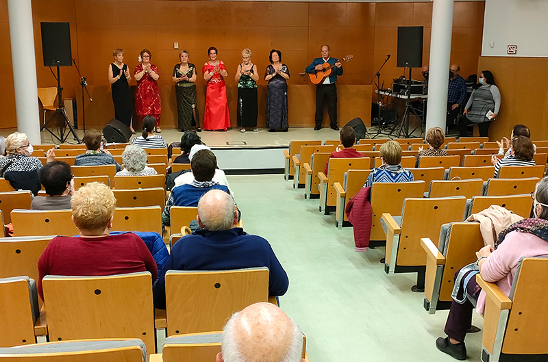 Santa Coloma de Gramenet. PlaybackMENT. View from the audience of elderly people watching a show. On stage, six women wearing long dresses clap their hands and a man plays the guitar.